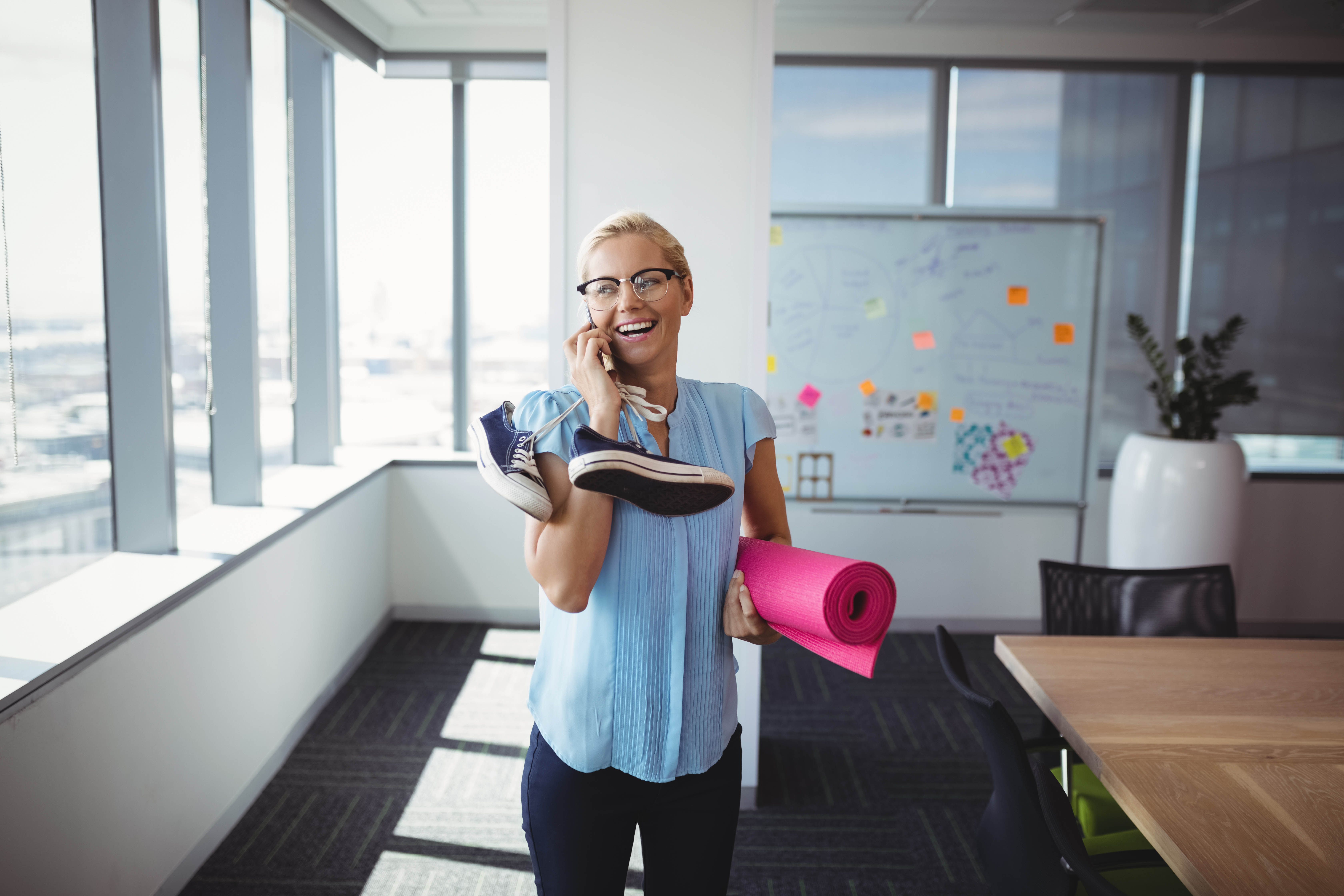 Young blond woman talks on the phone while carrying her yoga mat and running shoes in the office